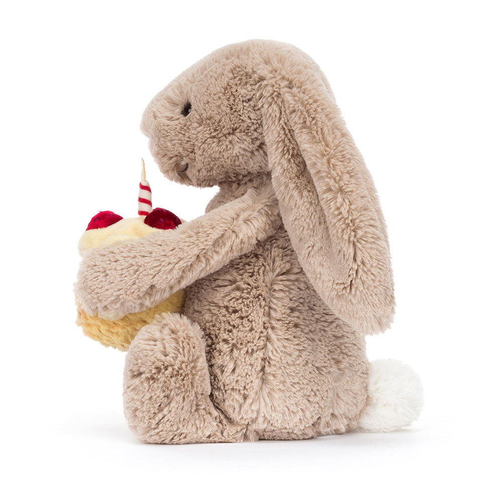 Seitenansicht: Jellycat Bashful Beige Bunny Birthday – sitzender beiger Kuscheltier-Hase mit Hängeohren und kleinem Geburtstagskuchen und Kerze in den Pfoten.