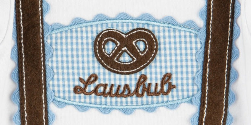 Detailaufnahme eines Babybodys mit gestickter Brezel, dem Schriftzug ‚Lausbub‘ auf einer blau-weiß karierten Vichy-Applikation und angedeuteten braunen Hosenträgern.