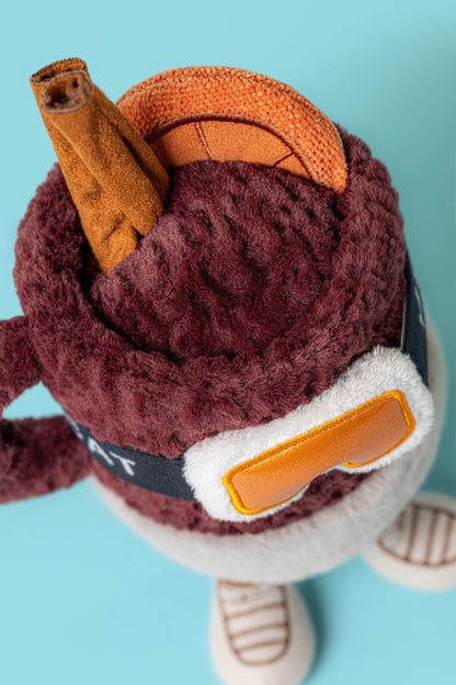 Detailansicht von oben des Jellycat Amuseables Mullover Spiced Drink mit Füßchen, Skibrille, Ohrenschützern, Zimtstange, Orangenscheibe