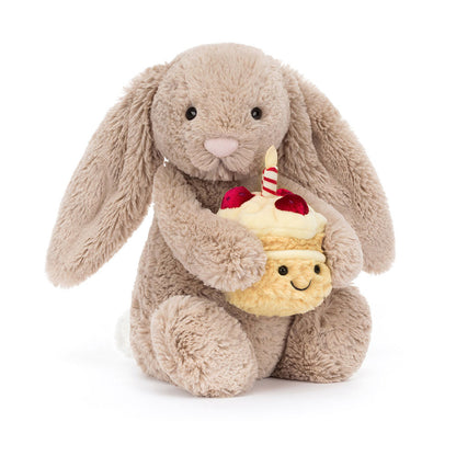 Vorderansicht: Jellycat Bashful Beige Bunny Birthday – sitzender beiger Kuscheltier-Hase mit Hängeohren und kleinem Geburtstagskuchen und Kerze in den Pfoten.