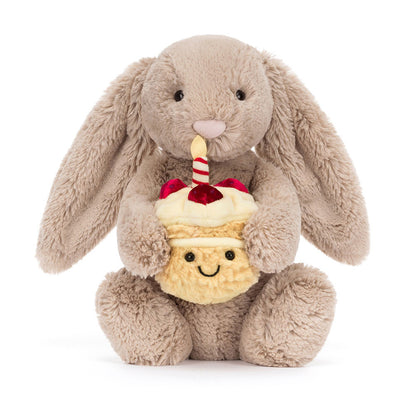 Vorderansicht: Jellycat Bashful Beige Bunny Birthday – sitzender beiger Kuscheltier-Hase mit Hängeohren und kleinem Geburtstagskuchen und Kerze in den Pfoten.