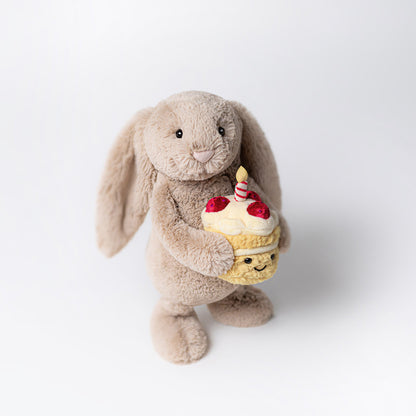 Vorderansicht: Jellycat Bashful Beige Bunny Birthday – sitzender beiger Kuscheltier-Hase mit Hängeohren und kleinem Geburtstagskuchen und Kerze in den Pfoten.