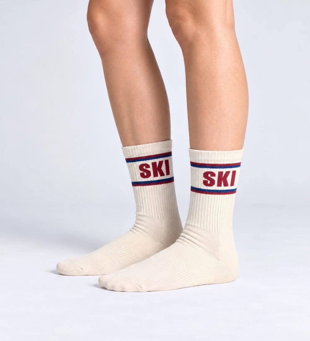 Cremefarbene Unisex-Socken SKI von No Bad Days Club mit rotem Schriftzug und blau-roten Streifen, an den Füßen getragen.