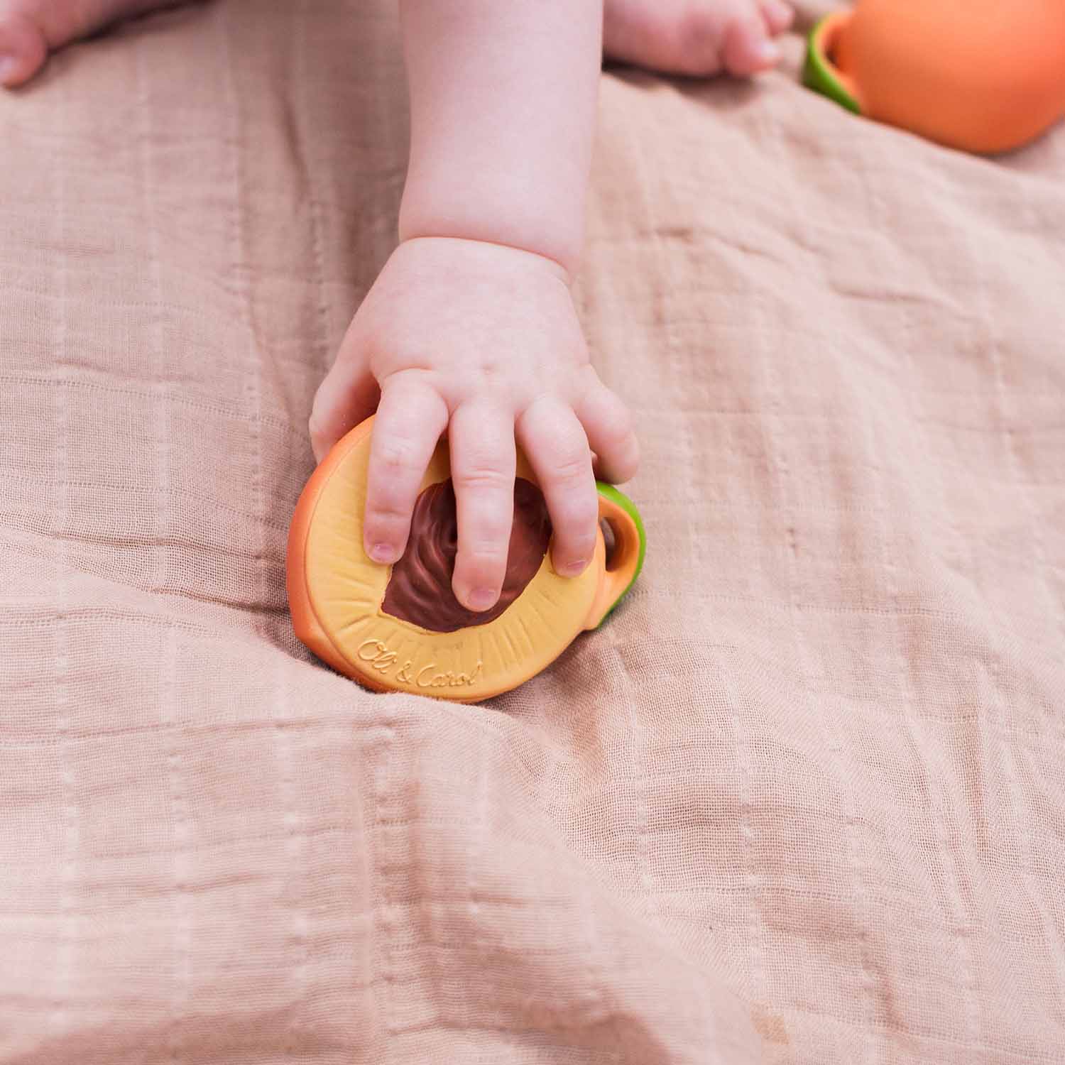 Baby hält den Palm Peach Beißring aus Naturkautschuk in den Händen