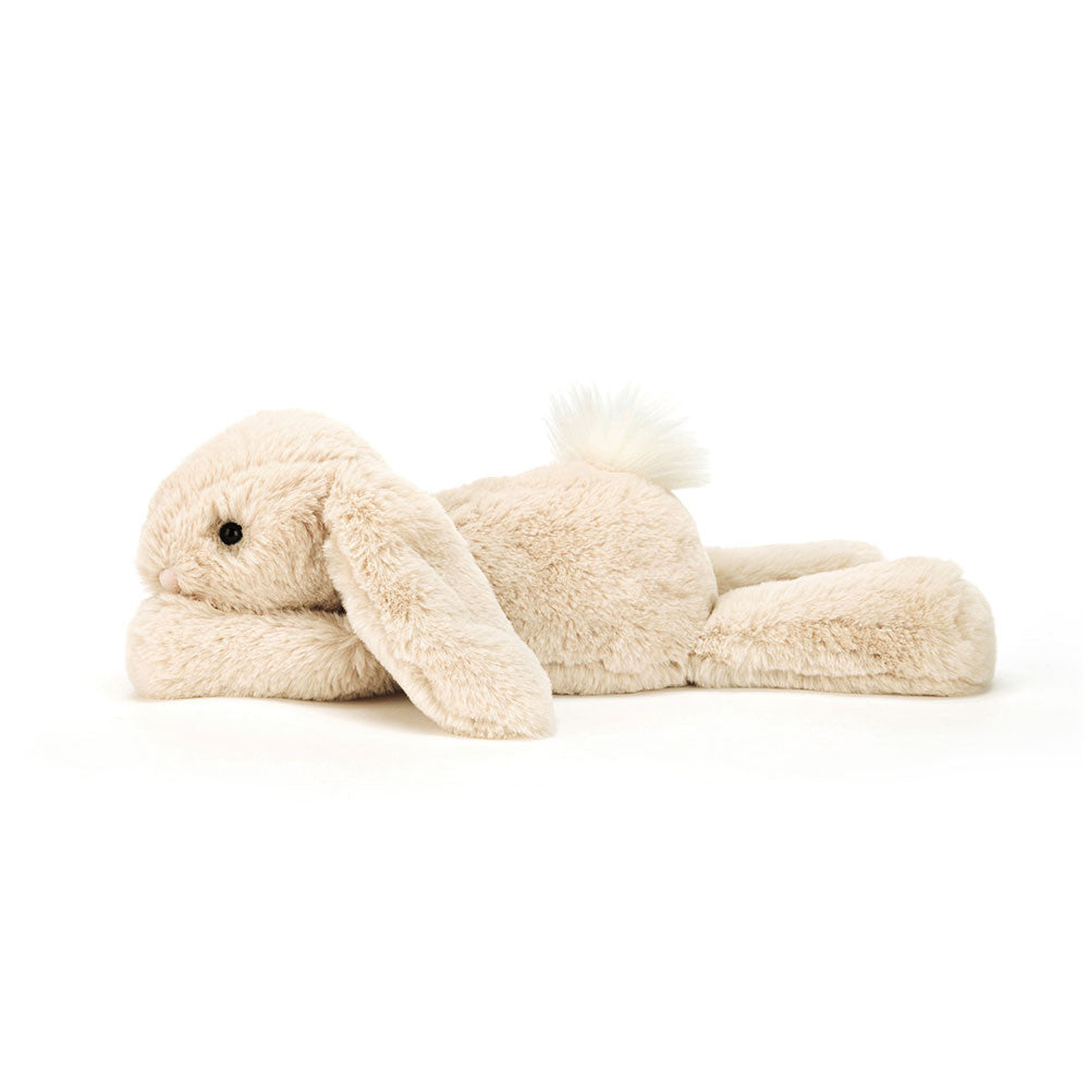 Seitenansicht: Jellycat Smudge Rabbit – liegender hellbeiger Kuschelhase mit langen Hängeohren, ausgestreckten Beinen und Kopf auf den Vorderpfoten. Mit kleinen schwarzen Knopfaugen und weißem Puschelschwanz.