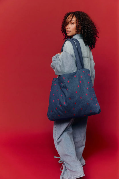 Frau trägt eine Studio Noos Mom-Bag über der Schulter aus dunkelblauem Denim mit wattierten Henkeln, gestickten roten Herzen und Logo