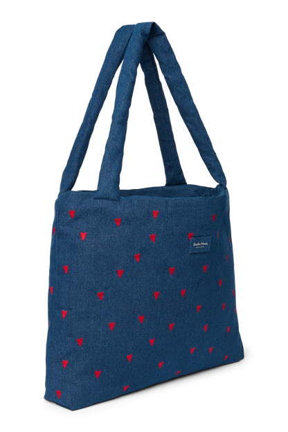 Schräge Ansicht der Studio Noos Mom-Bag aus dunkelblauem Denim mit wattierten Henkeln, gestickten roten Herzen und Logo