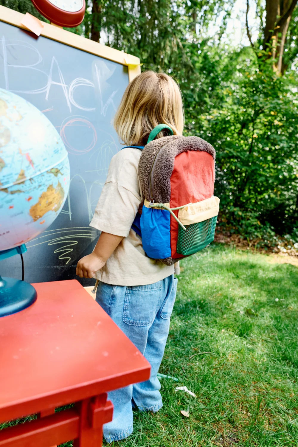Junge mit Studio Noos Patchwork Mini-Rucksack Fiery Red in bunten Farben, braunem Teddy-Stoff, roten Patchwork-Details und blau-grün-gelben Akzenten an einer Schreibtafel
