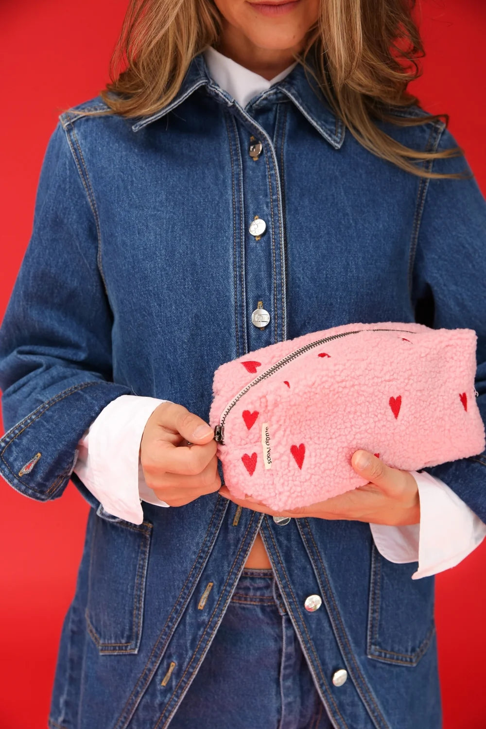 Frau hält Studio Noos Pouch aus pinkem Teddystoff mit gestickten roten Herzen, Reißverschluss und Studio Noos Label in den Händen