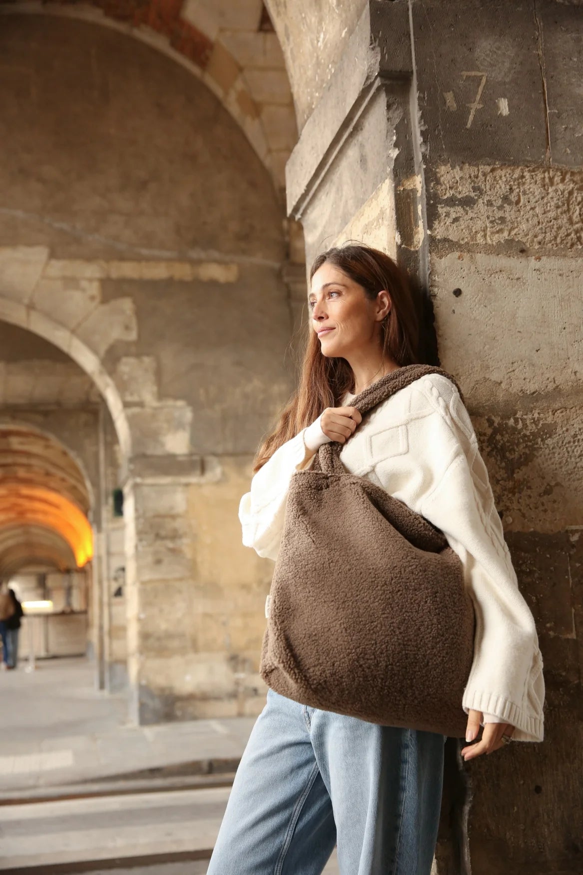 Foto: Eine Frau lehnt an einer Wand und trägt eine Studio Noos Teddy Mom-Bag l Braun aus weichem braunem Teddy-Stoff mit breiten Henkeln